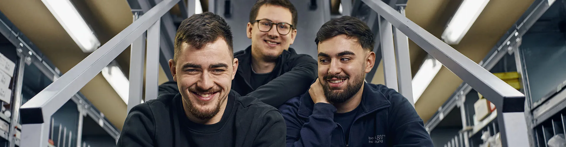 Eine Gruppe von drei jungen Handwerkern sitzt auf einer Metalltreppe in einem modernen Werkstattbereich.
