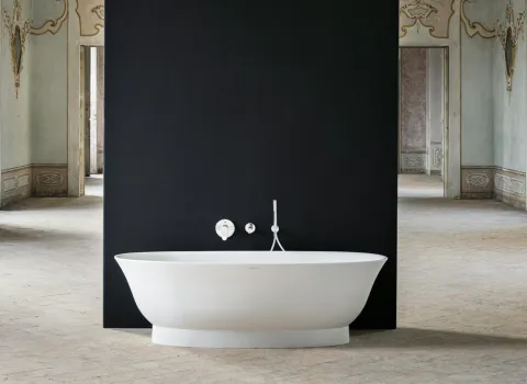 Eine elegante, freistehende weiße Badewanne steht vor einer mattschwarzen Wand in einem großen, historischen Raum mit dekorativen Stuckverzierungen.