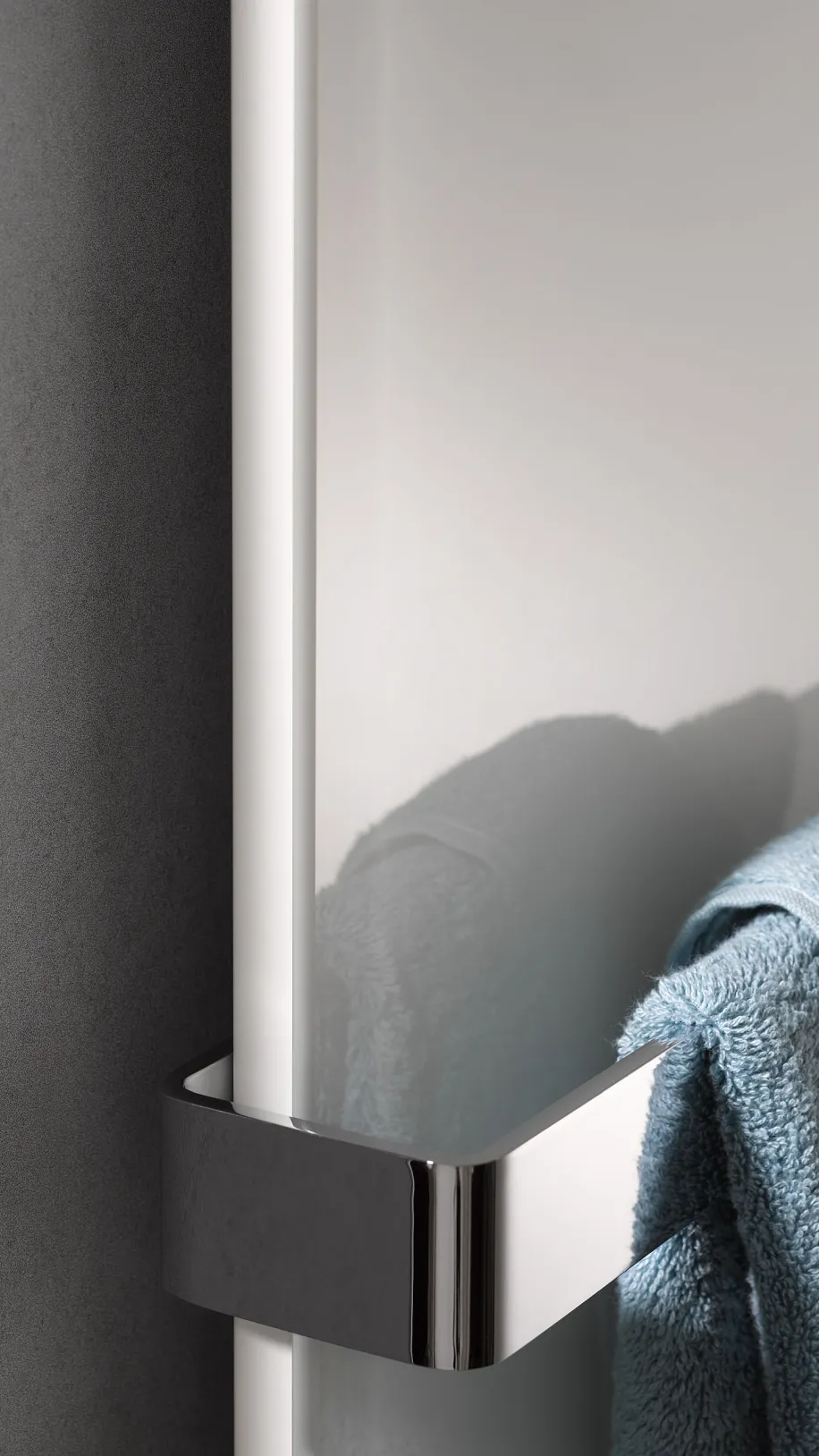 Detailaufnahme eines Glasheizkörpers mit verchromter Halterung, an der ein blaues Handtuch hängt, vor einer dunklen Wand.