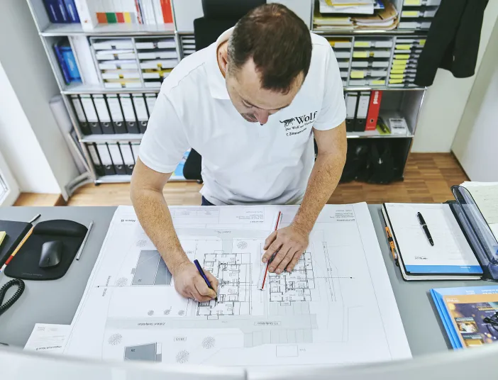 Ein technischer Systemplaner der Firma bad und heizung Wolf steht an einem Schreibtisch und arbeitet konzentriert an einem Bauplan mit Maßstab und Bleistift.